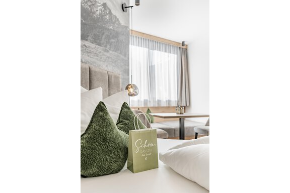 A modern hotel room with cozy, green cushions and bright, minimalist decor. A small gift bag labeled "Nice to have you here" lies on the bed. - Marco's Mountain Home - Sölden