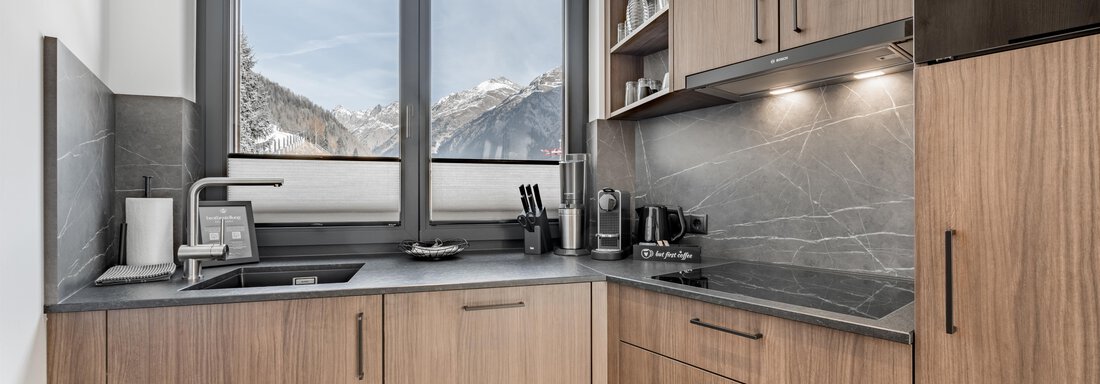 Modern kitchen with wooden cabinets and gray countertops. A mountain landscape is visible through a large window. - Marco's Mountain Home - Sölden