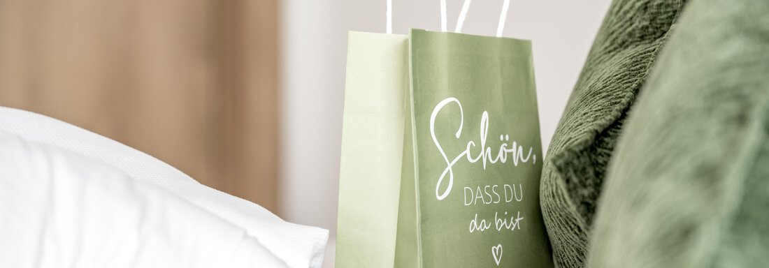 A green paper bag stands on a bed next to pillows. The bag has the inscription "Nice to have you here". - Marco's Mountain Home - Sölden