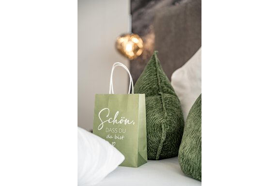 A green paper bag with the inscription "Nice to see you" stands on a bed next to two green, cushion-like decorative items. In the background, soft lighting is visible. - Marco's Mountain Home - Sölden