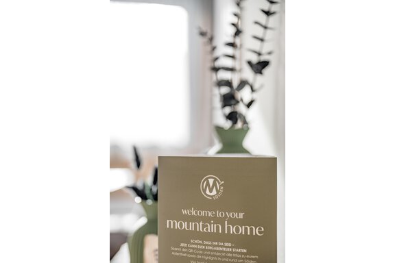 A welcome sign with the inscription "Welcome to Your Mountain Home" stands in front of a green vase with plants. The background is bright and cheerful. - Marco's Mountain Home - Sölden