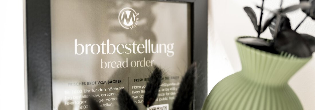 A frame with the inscription "Brotbestellung" and "bread order" is attached to the wall. Next to it stands a green vase with plants. - Marco's Mountain Home - Sölden