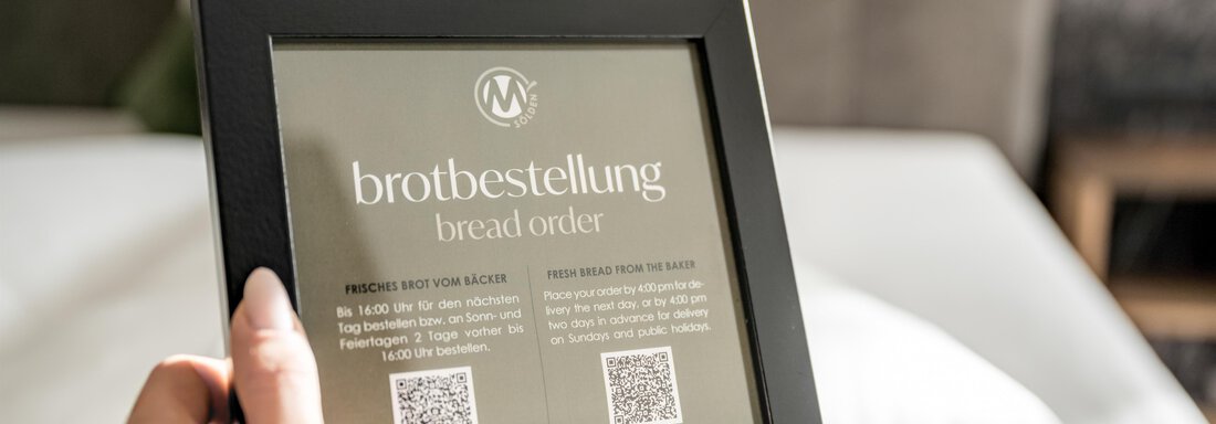 A handheld board for bread serving with QR codes and information. In the background, a cozy bed with pillows is visible. - Marco's Mountain Home - Sölden