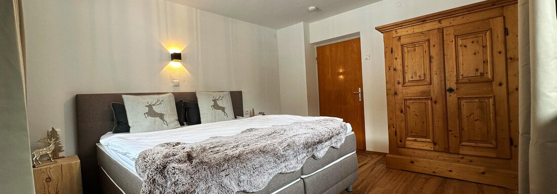 A cozy bedroom with a large bed, wooden wardrobe, and warm lighting. The floor is made of wood, and the walls are painted in light colors. - Mountain Lodge Sölden - Sölden