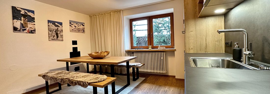A modern dining room with a large wooden table and benches. Pictures hang on the wall, and the kitchen has an open design. - Mountain Lodge Sölden - Sölden