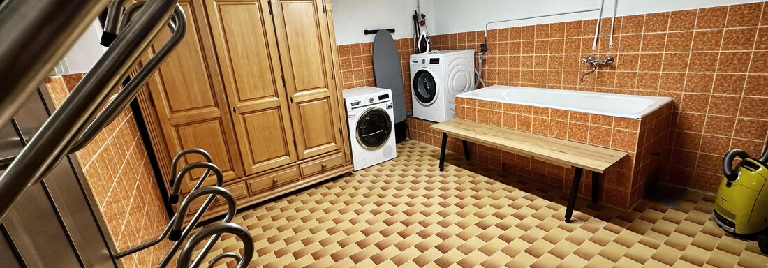 A functional laundry room with a washing machine, a dryer, and a wooden bench. The walls are tiled in orange and the floor has a geometric pattern. - Mountain Lodge Sölden - Sölden