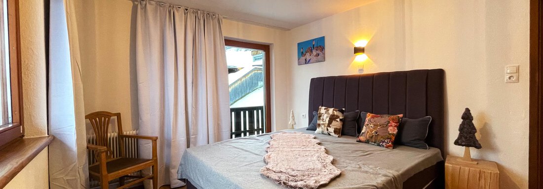 A cozy bedroom with a large bed and warm lighting. The room features a chair and a wooden table, as well as windows with curtains. - Mountain Lodge Sölden - Sölden