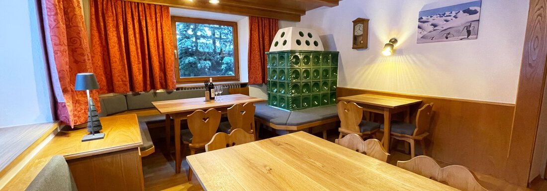 A cozy interior with wooden ceilings and large tables. The windows are decorated with orange curtains, and a green glass cabinet stands in the room. - Mountain Lodge Sölden - Sölden