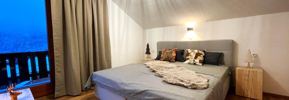 A modern bedroom with a large bed and warm wood elements. In front of the window, a view of a wintry landscape is visible. - Mountain Lodge Sölden - Sölden