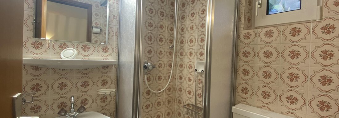 A small bathroom with a shower, sink, and toilet. The walls are decorated with a vintage tile pattern. - Mountain Lodge Sölden - Sölden
