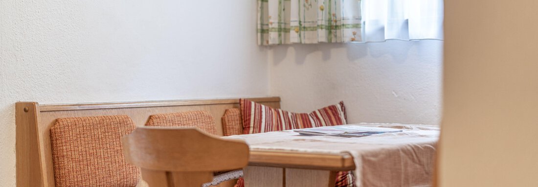 A cozy dining area with a wooden table and upholstered benches. A window with curtains lets in soft light. - Pension Birkenheim - Sölden