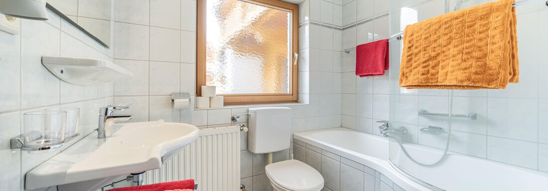 A modern bathroom with a shower and bathtub. The walls are tiled in white and colored towels add a vibrant accent. - Pension Birkenheim - Sölden