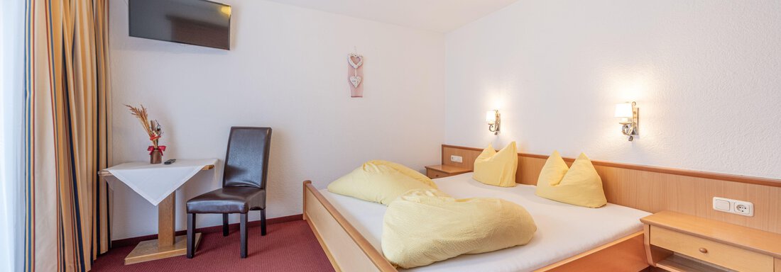A cozy room with a double bed, two large cushions, and a wall-mounted TV. A small seating area with a table and a chair completes the furnishings. - Pension Birkenheim - Sölden