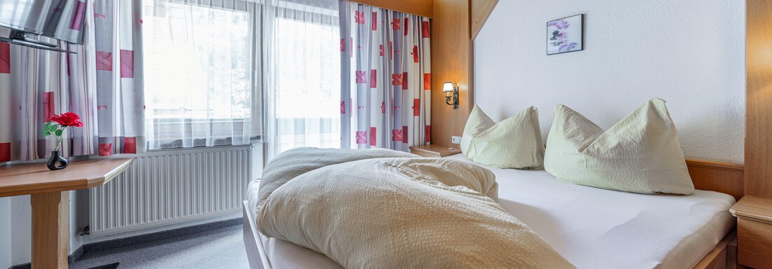 A cozy bedroom with a large bed and pillows. A table and a television are also present in the room, with windows that let in daylight. - Pension Birkenheim - Sölden