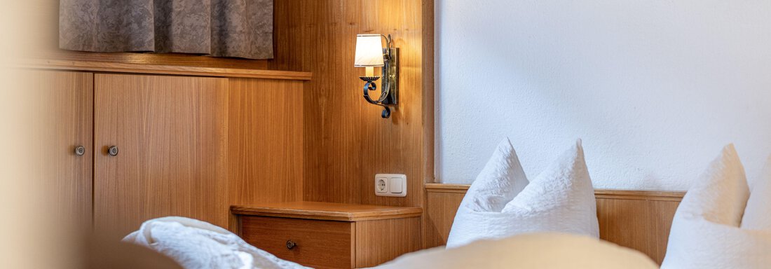 A cozy bedroom with wood paneling and warm light. The bed is decorated with pillows and blankets. - Pension Birkenheim - Sölden