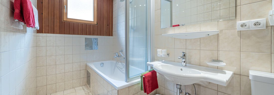 A modern bathroom with a bathtub, a shower, and a sink. White tiles and wood paneling create a bright, friendly atmosphere. - Pension Birkenheim - Sölden
