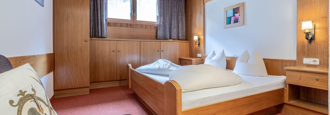 A cozy bedroom with a wooden bed and a comfortably looking sofa. The room is bright and friendly, with a large window that lets in daylight. - Pension Birkenheim - Sölden