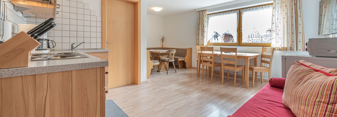 A modern kitchen with wooden cabinets and an open dining area. The room is bright and friendly with large windows and a red sofa. - Pension Birkenheim - Sölden