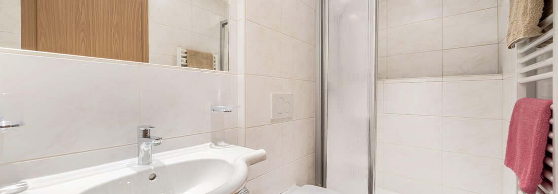 A modern bathroom with a shower, a sink, and a toilet. The walls are light-tiled and the furnishings are functional. - Pension Birkenheim - Sölden