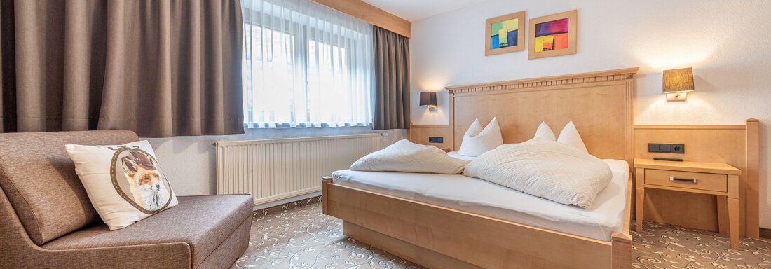 A cozy hotel room with a double bed and a comfortable armchair.  
Brightly lit windows and subtle colors create an inviting atmosphere. - Pension Birkenheim - Sölden