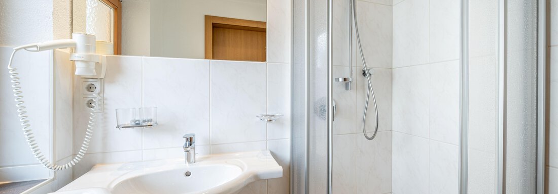 A modern bathroom with a shower and a washbasin. It is bright and clean, with white tiles and a large window. - Pension Birkenheim - Sölden