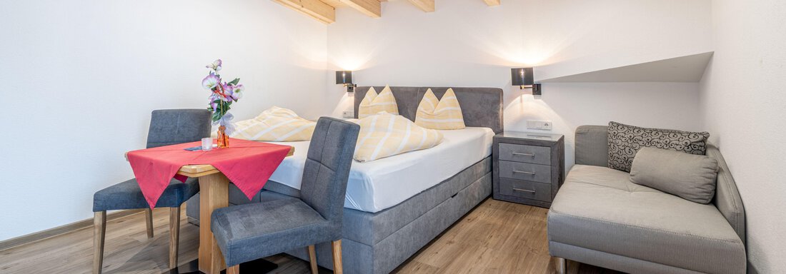 A cozy room with a double bed, a sofa, and a dining table. The wood decor and warm colors create an inviting atmosphere. - Pension Birkenheim - Sölden