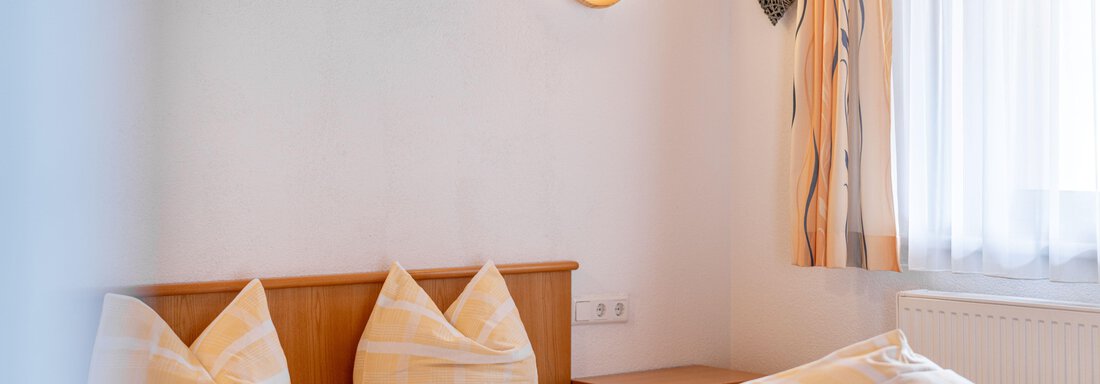 A cozy bedroom with a wooden bed and pillows in yellow and white stripes. The room has bright walls and a window with curtains that lets in daylight. - Pension Birkenheim - Sölden