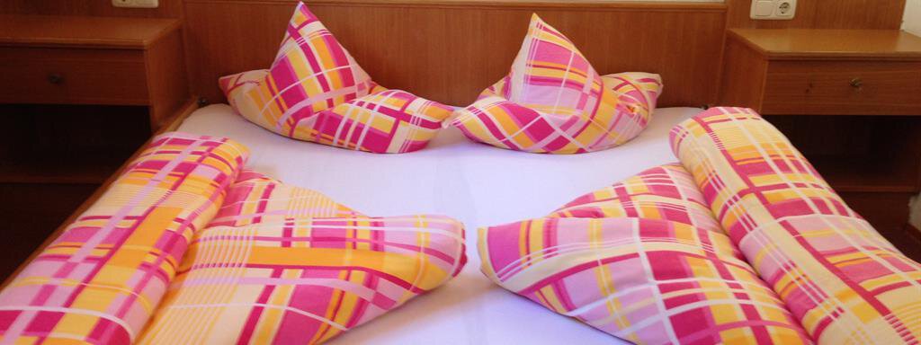 A cozy bedroom with a double bed. The bed linens have a striking striped pattern in pink and yellow. - Pension Birkenheim - Sölden