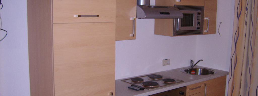 Küchenblock A modern kitchen with wooden cabinets and stainless steel appliances. It includes a stove, a refrigerator, and a microwave. - Pension Birkenheim - Sölden