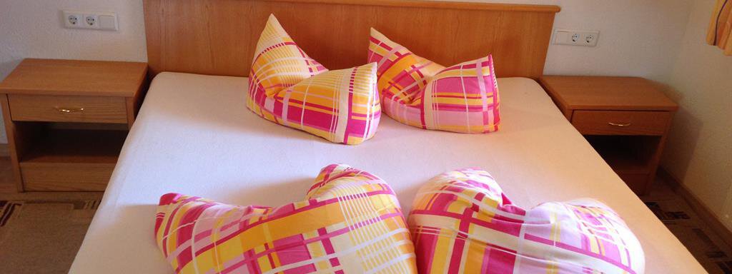 A cozy bedroom with a bed and two nightstands. The pillows are heart-shaped and designed with colorful check patterns. - Pension Birkenheim - Sölden