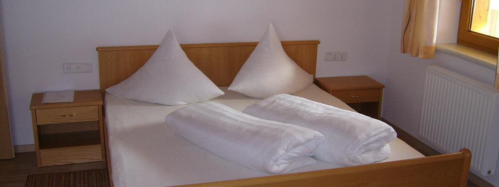 A simple bedroom with a double bed and two placed pillows. The room has a wooden floor and a window with curtains. - Pension Birkenheim - Sölden
