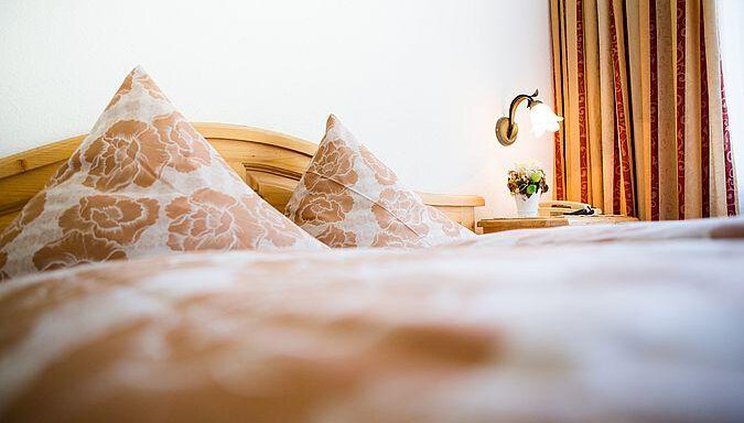 A cozy bedroom with a wooden bed and decorative pillows. Next to the bed is a small lamp and a floral decoration. - Pension Charly - Sölden
