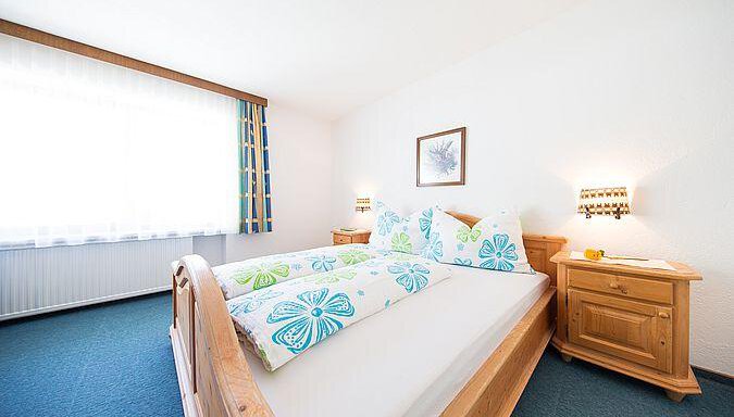 A bright bedroom with a large bed and colorful pillows. There is a nightstand and a window with curtains. - Pension Charly - Sölden