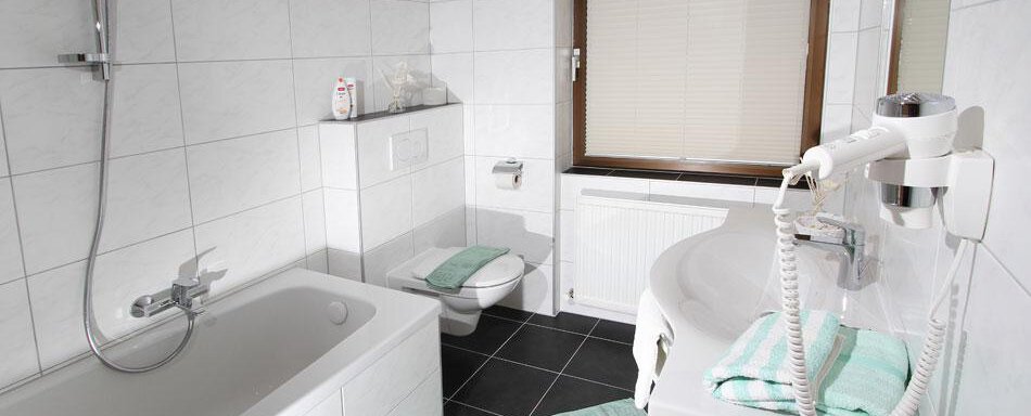 A modern bathroom with white wall tiles and a dark floor. It features a bathtub, a sink, and a toilet as well as green towels. - Pension Charly - Sölden