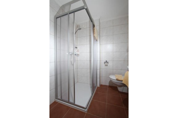 A modern bathroom with a glass shower and a toilet. The walls are light tiled and the floor is brown. - Pension Charly - Sölden