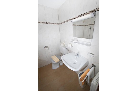 A modern bathroom with white tiles and a large mirror. There is a toilet, a sink, and fresh towels. - Pension Charly - Sölden