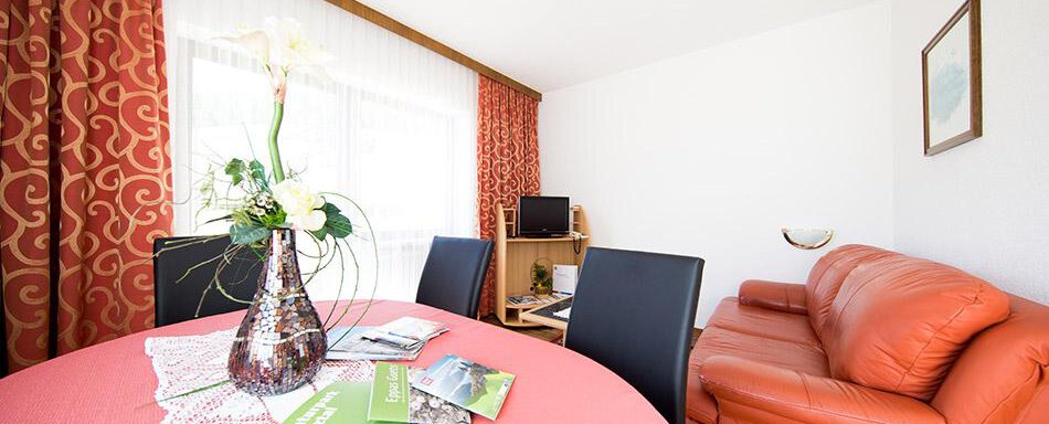 Wohnraum mit Essbereich A cozy living space with a round table and an orange sofa. On the table stands a vase with a flower and some informational brochures. - Pension Charly - Sölden