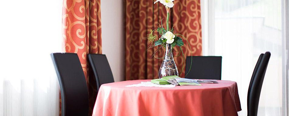 A small table with a red tablecloth and a vase with flowers. Two black chairs sit around the table, and in the background, curtains are visible. - Pension Charly - Sölden
