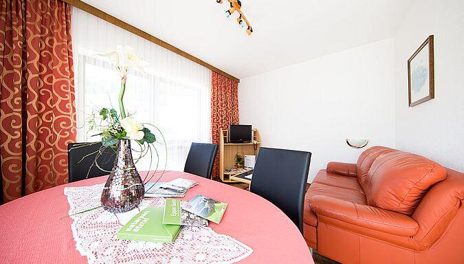 A cozy living space with a red sofa and a dining table. On the table, there is a flower vase and some brochures. - Pension Charly - Sölden