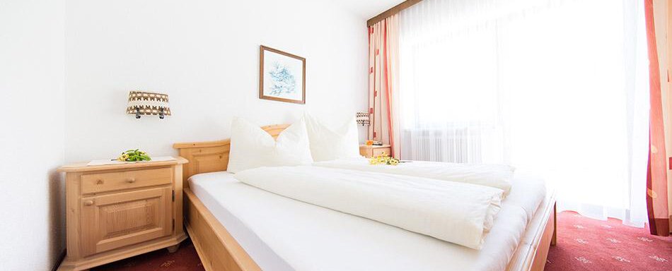 A bright bedroom with two beds and large windows. The furnishings are made of wood and the floor is covered with red carpet. - Pension Charly - Sölden
