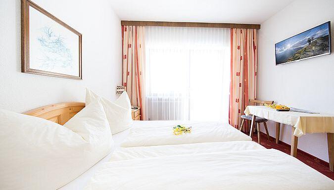 A bright room with two single beds and large windows. A table with a small flower arrangement completes the pleasant atmosphere. - Pension Charly - Sölden