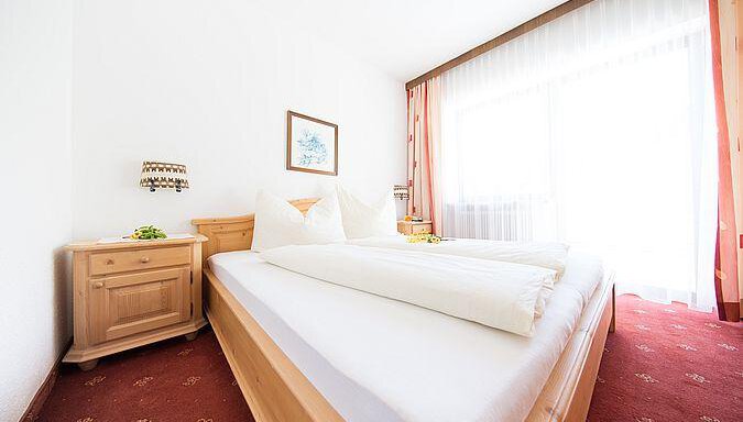 A bright bedroom with two beds and a large window. The room is cozy with wooden furniture and red carpet flooring. - Pension Charly - Sölden