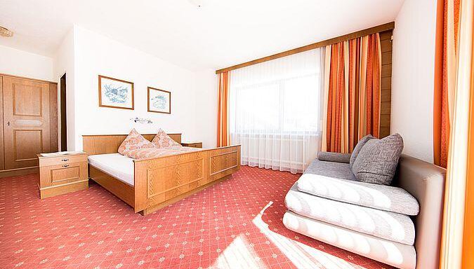 A bright room with a large bed and a cozy seating area. The windows let in plenty of daylight, and the orange curtains give the room a warm atmosphere. - Pension Charly - Sölden