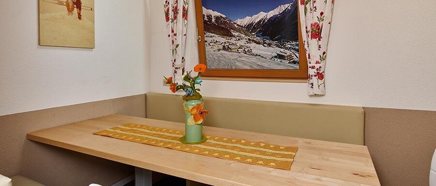 Essbereich A cozy dining area with a simple wooden table and white chairs. On the wall, there is a window showcasing a mountain landscape as well as a vase with colorful flowers. - Pension Gritsch Ewald - Sölden