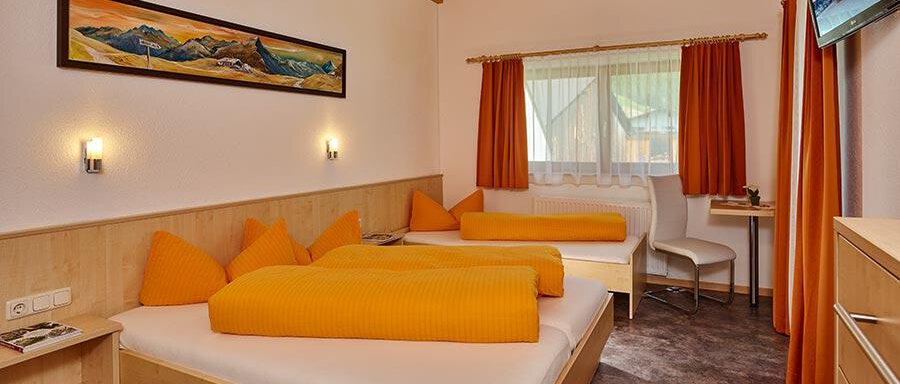 A cozy bedroom with two beds and orange pillows. The walls are light, and there is a window with curtains as well as a small table. - Pension Gritsch Ewald - Sölden