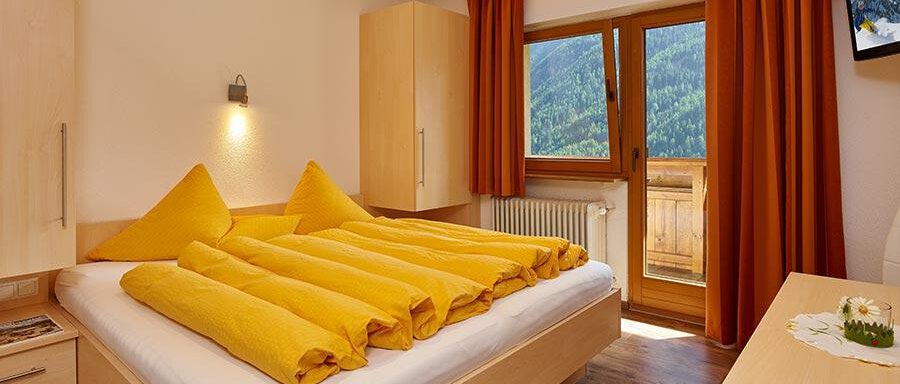 A cozy bedroom with a large bed and bright yellow blankets. The room has large windows overlooking the mountains and is brightly designed. - Pension Gritsch Ewald - Sölden