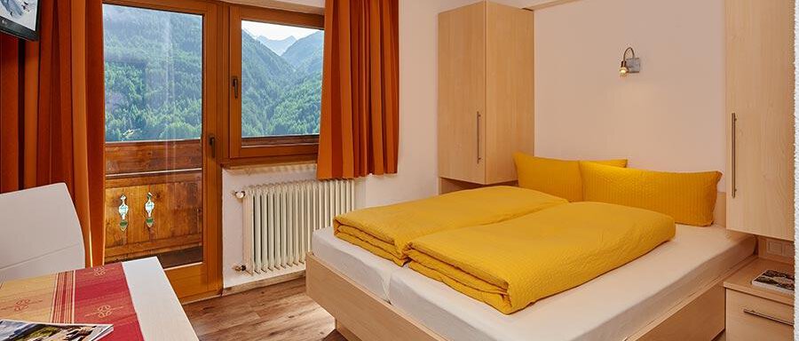 A cozy bedroom with a double bed and yellow blankets. Large windows offer a view of the surrounding mountains. - Pension Gritsch Ewald - Sölden