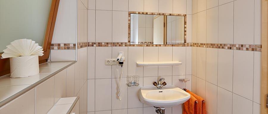 A modern bathroom with white tiles and a small sink. There is a towel holder with an orange towel and a large window with daylight. - Pension Gritsch Ewald - Sölden