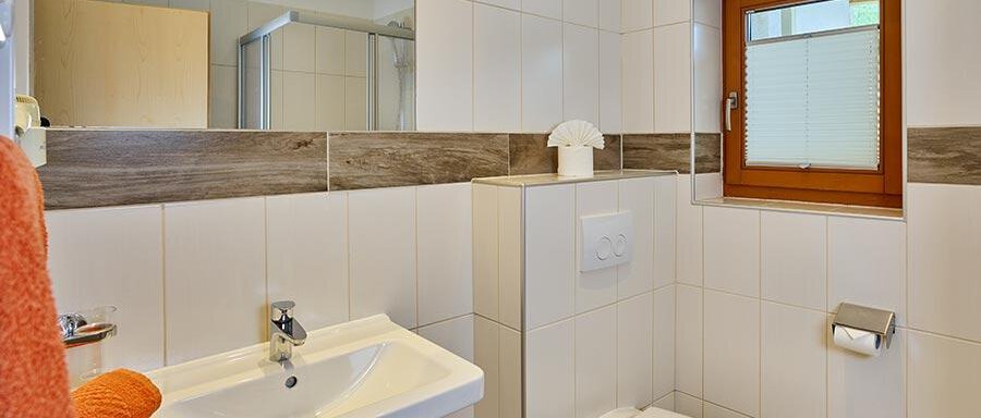 A modern bathroom with white tiles and wooden accents. It features a sink, a toilet, and a window with daylight. - Pension Gritsch Ewald - Sölden