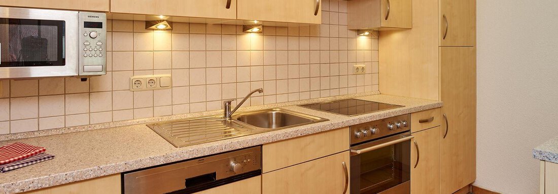 Küche A modern kitchen unit with wooden cabinets and bright countertops. It includes a sink, stove, oven, and microwave. - Pension Gritsch Ewald - Sölden